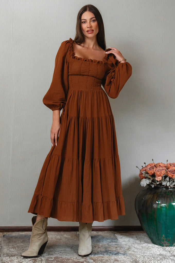 Woman wearing a brown dress with long sleeves standing against a plain wall with a vase of flowers in the background.