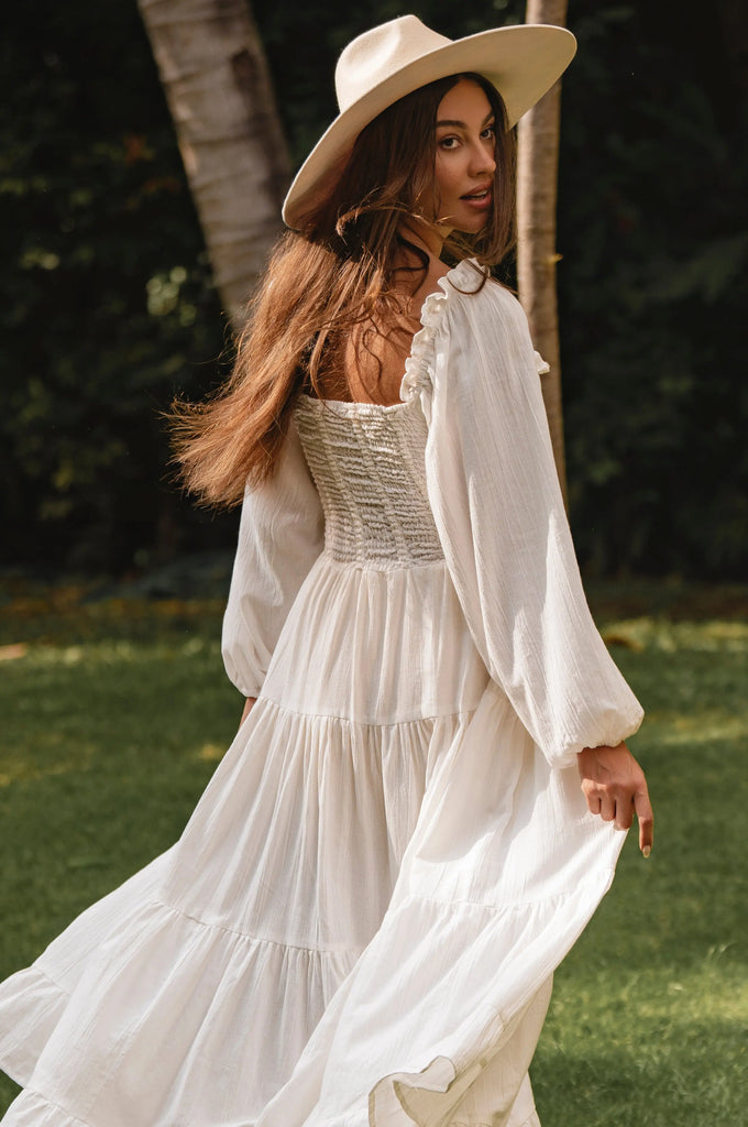Woman in a white dress and hat standing outdoors with greenery in the background cottage core style