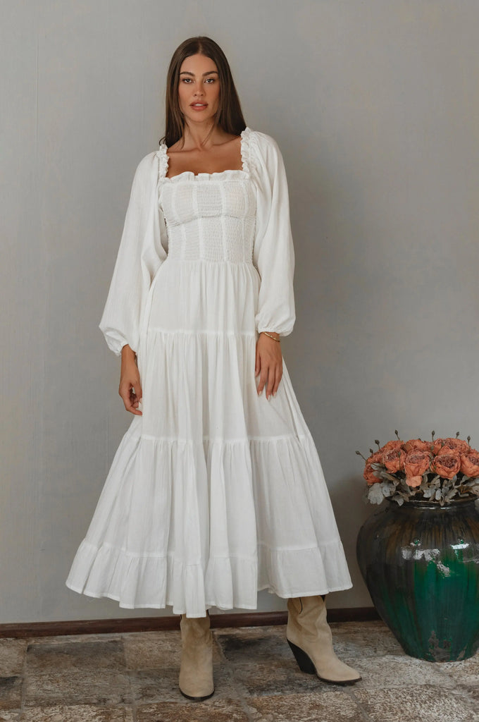 Woman wearing a white long sleeve dress standing next to a vase with flowers against a gray wall.