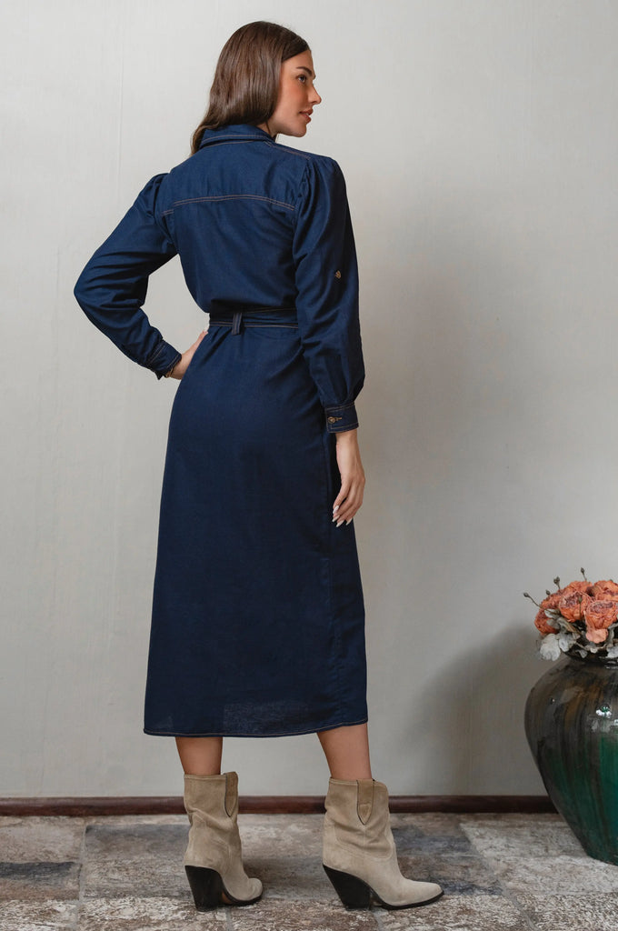 Woman wearing a long navy dress standing against a plain wall with a vase of flowers - cottage core style